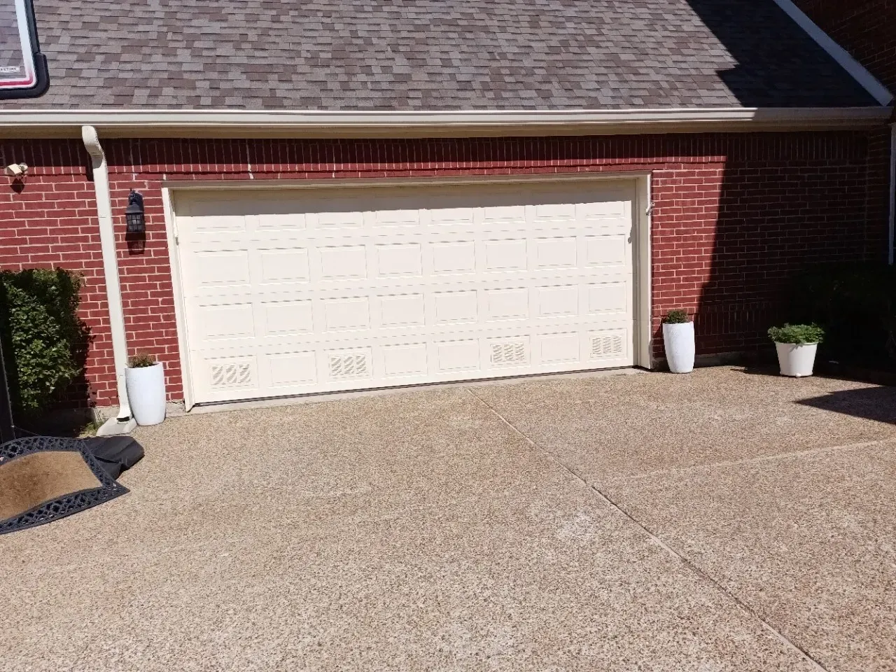 A tan garage door set in a red brick exterior with white potted plants on either side of the driveway.