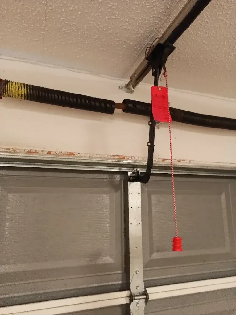 A close-up view of a garage door torsion spring and the manual emergency release rope with a red handle.