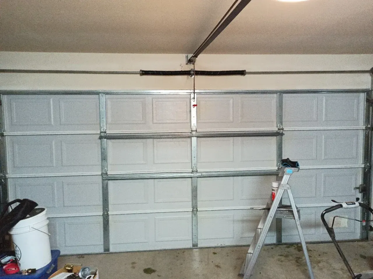 A garage door with horizontal panels, a metal spring mechanism across the top, a stepladder, and items on the floor.