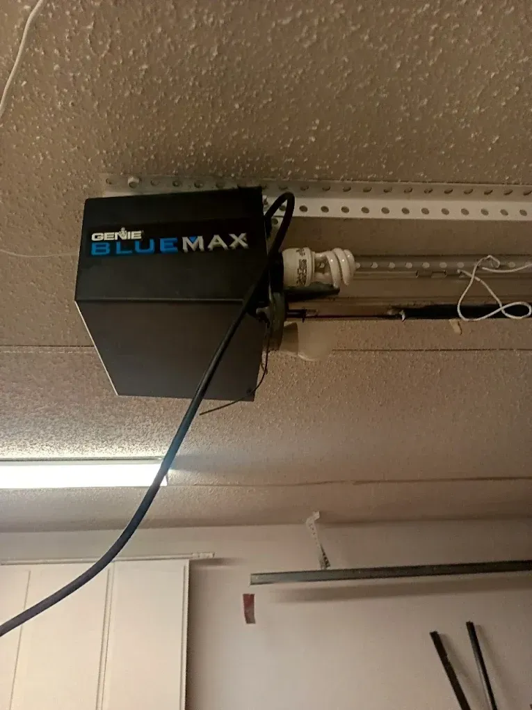 A black Genie Blue Max garage door opener mounted to a ceiling with a light bulb socket exposed on the side.