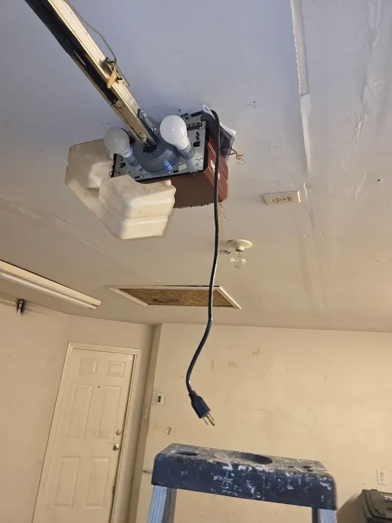 A garage door opener is mounted to a ceiling with a black power cord hanging down near a step ladder.