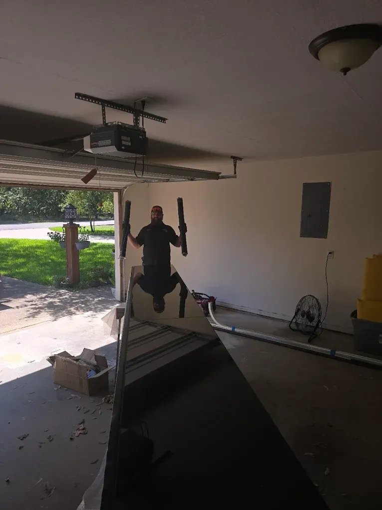 A person in a garage holds a large, reflective mirror, creating a clear reflection of themselves and the room.