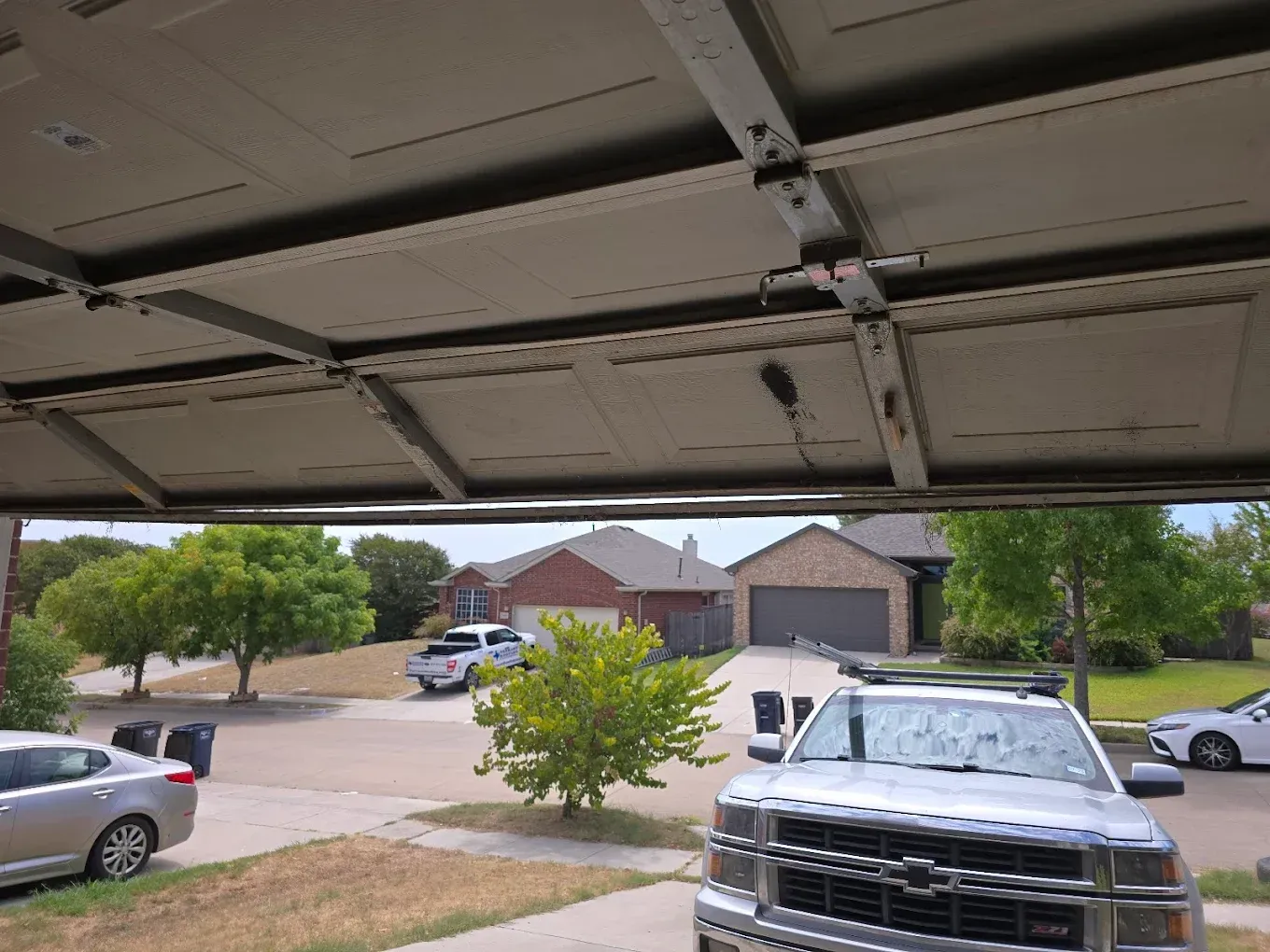 View from inside a garage looking out at a suburban street with parked cars, houses, and trees on a sunny day.