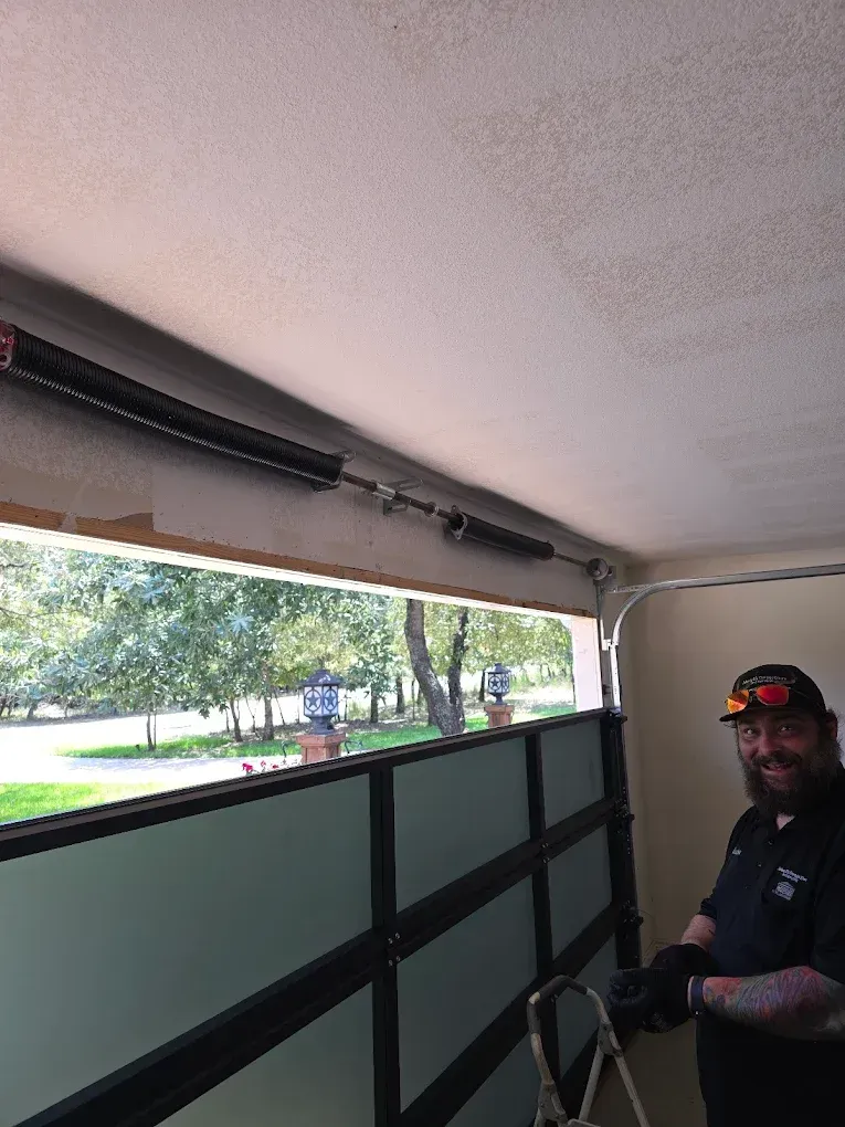 A smiling technician in a black uniform working on an overhead garage door torsion spring system.