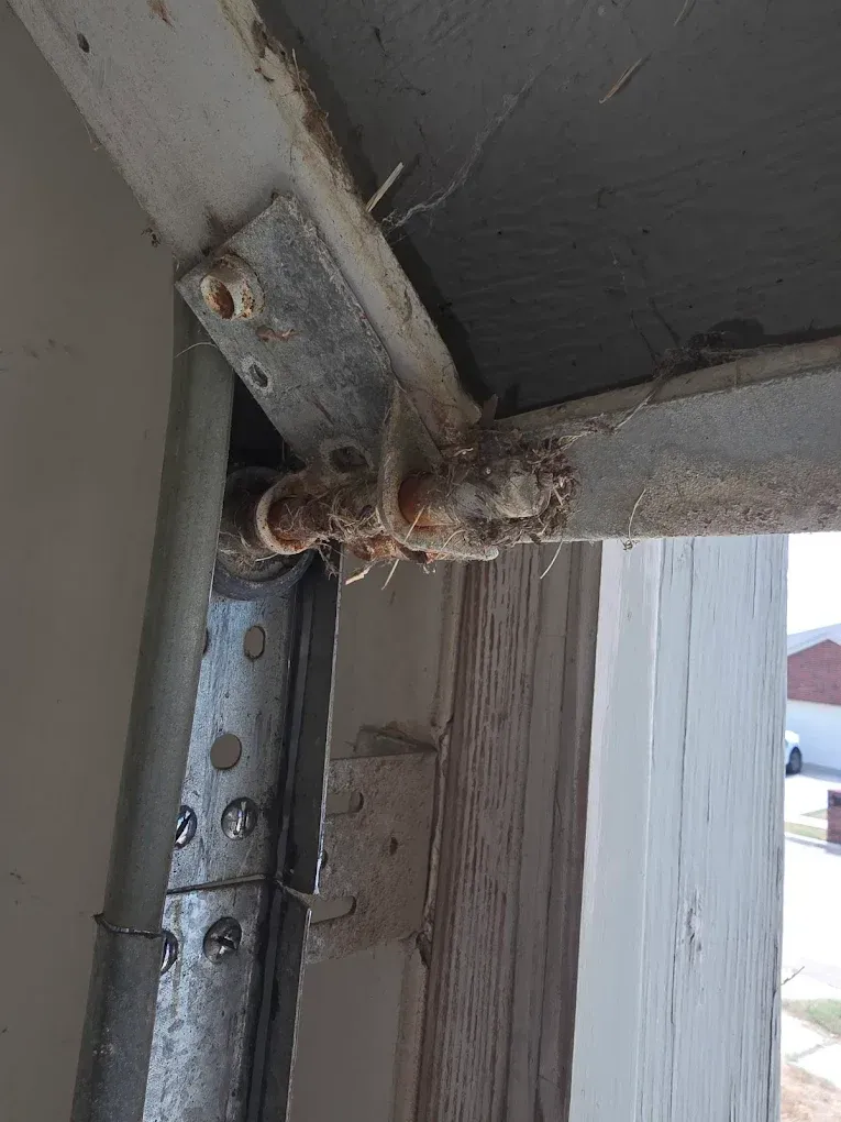 A close-up view showing a rusted, corroded metal hinge and roller assembly attached to a garage door track and header.
