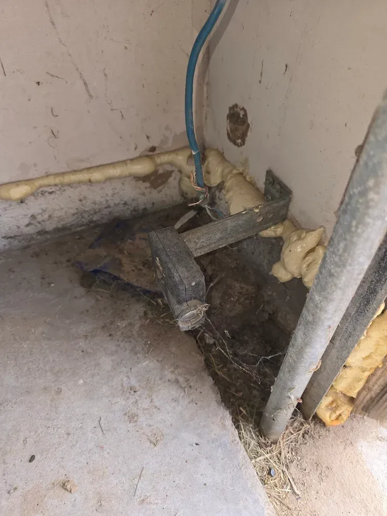 A corner of a room with a concrete floor, a metal bracket bolted to the wall, a blue pipe, and gaps filled with foam.