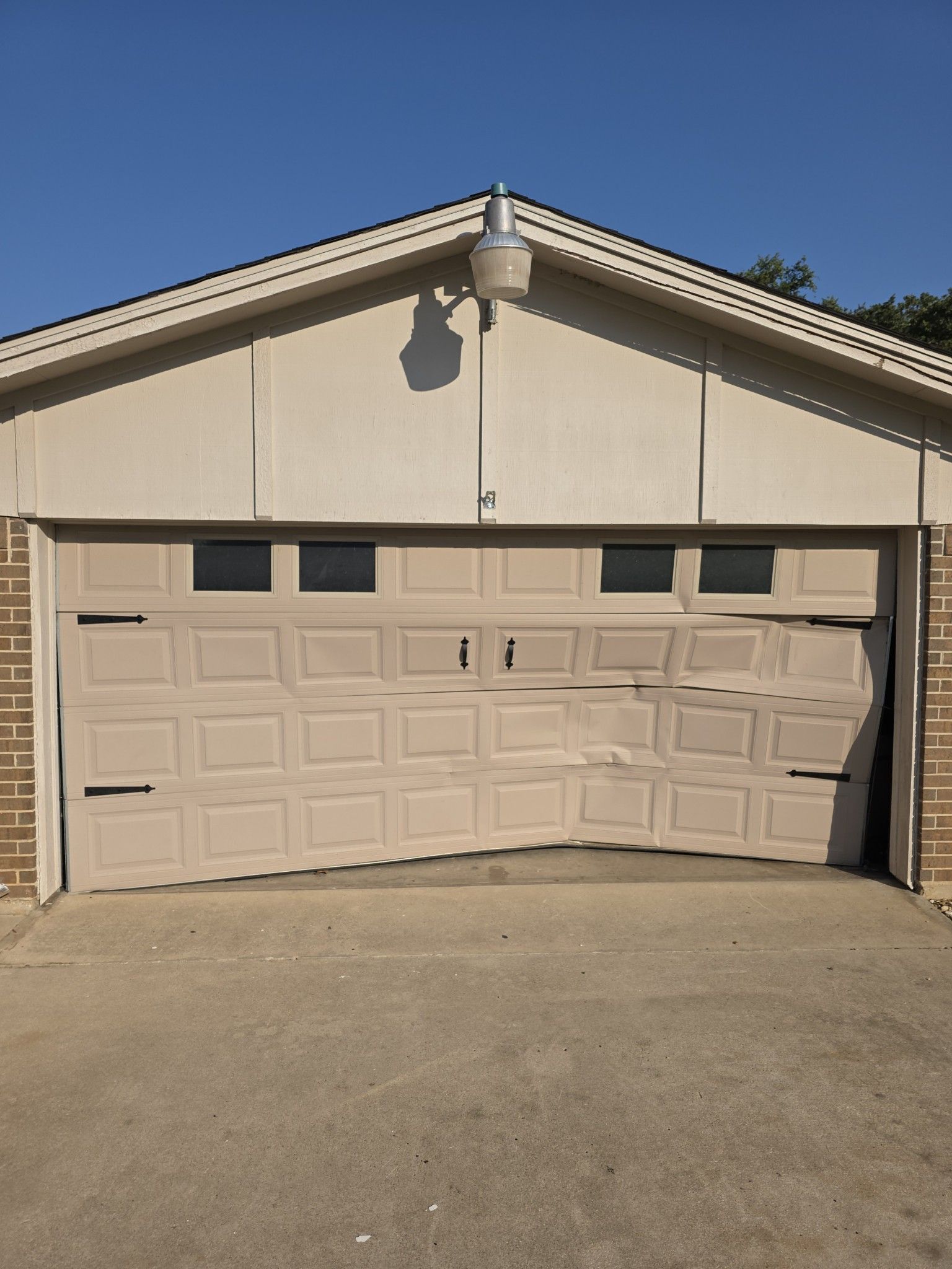 A beige garage door with significant structural damage, appearing buckled and bent in the center on a sunny day.
