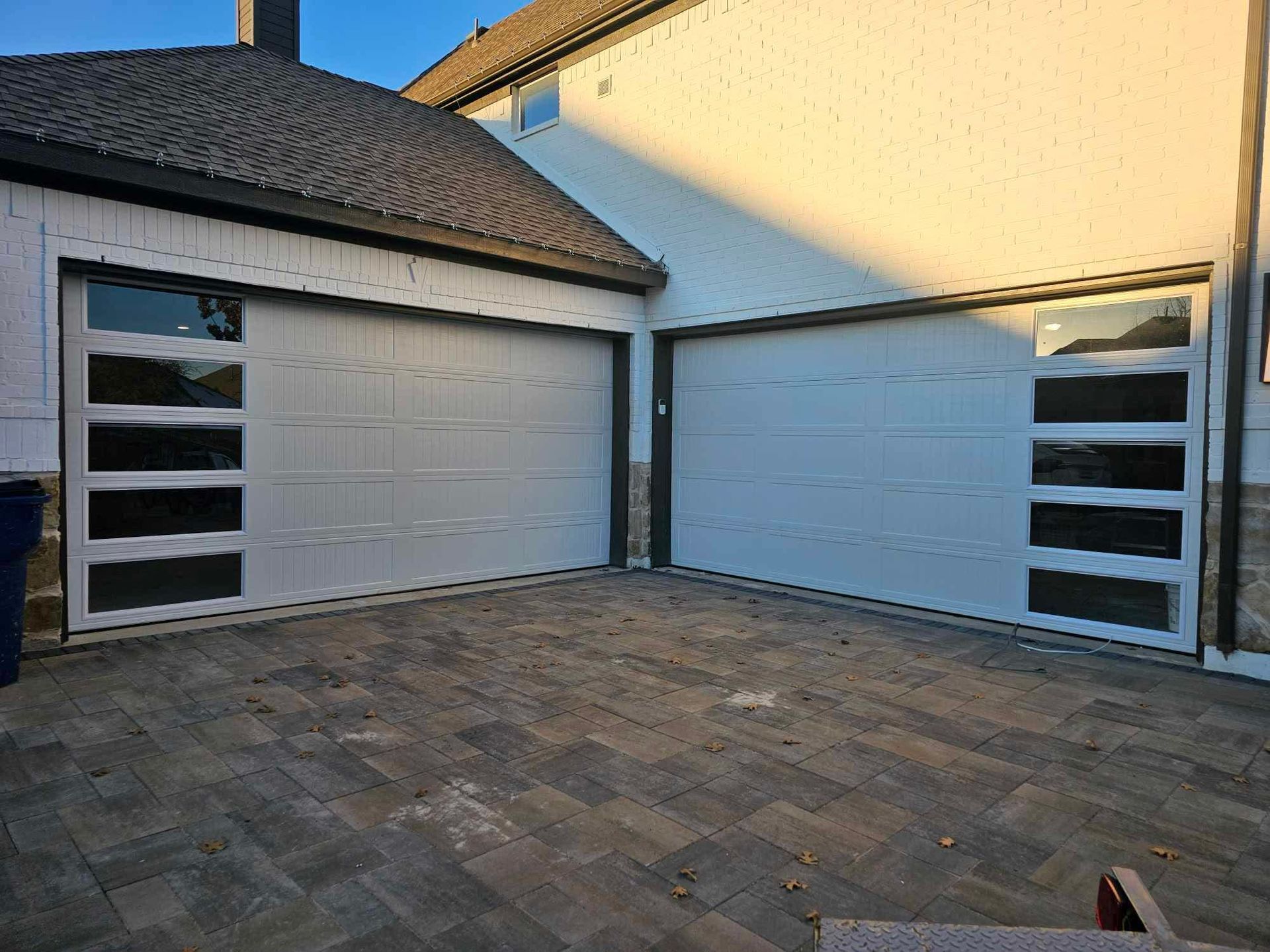 Two white garage doors with vertical windows on one side of each, set in a light-colored building with a paved driveway.
