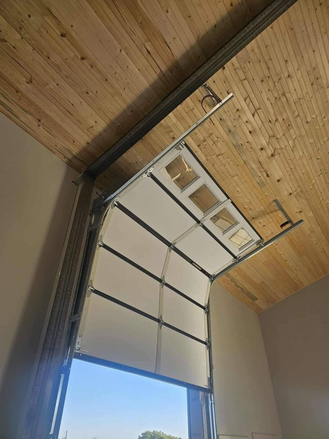 A sectional garage door with a window panel retracting along metal tracks into a high ceiling with wood paneling.
