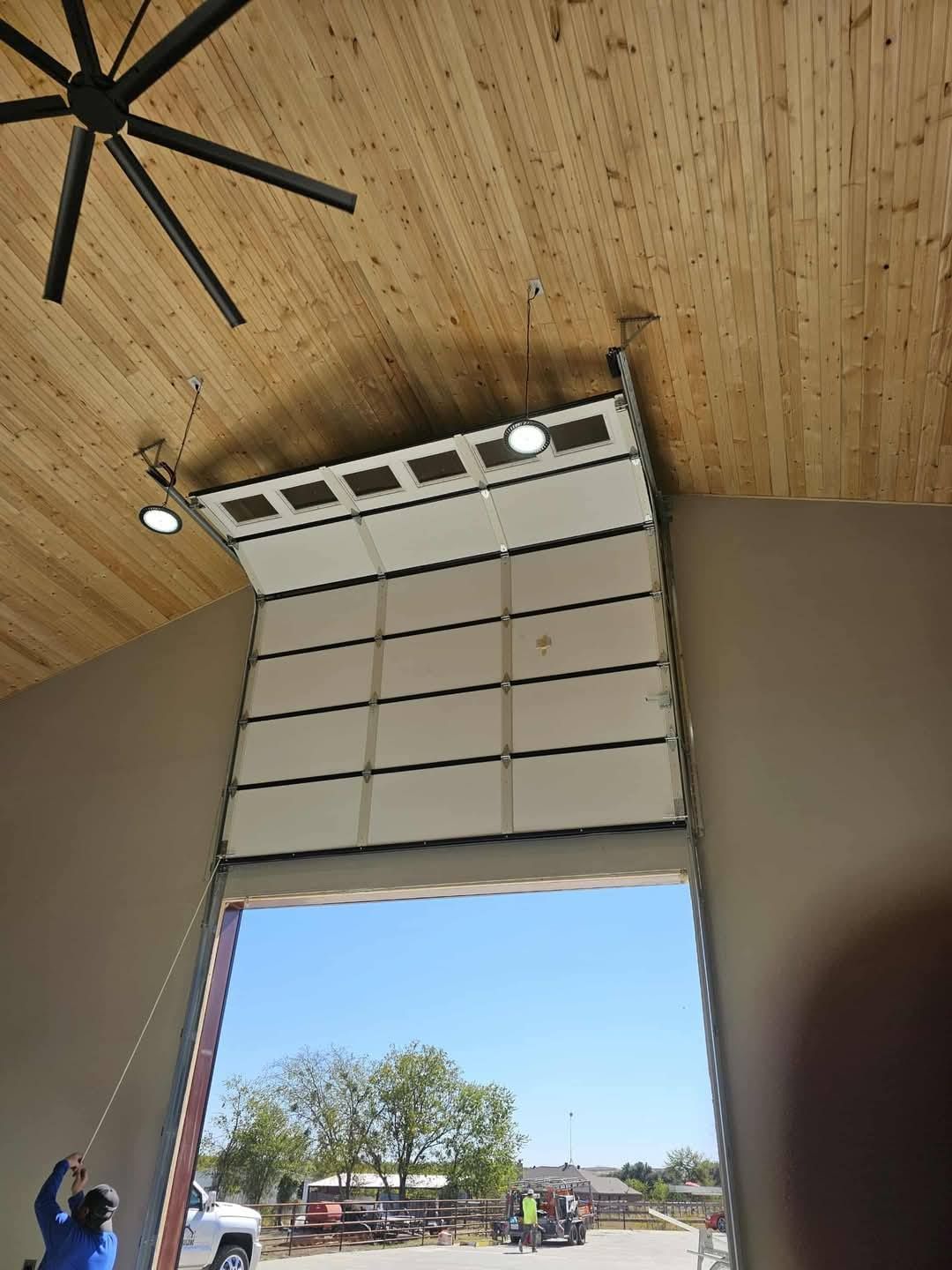 A person pulls a rope to open a white sectional garage door in a large workshop with a high wooden ceiling.