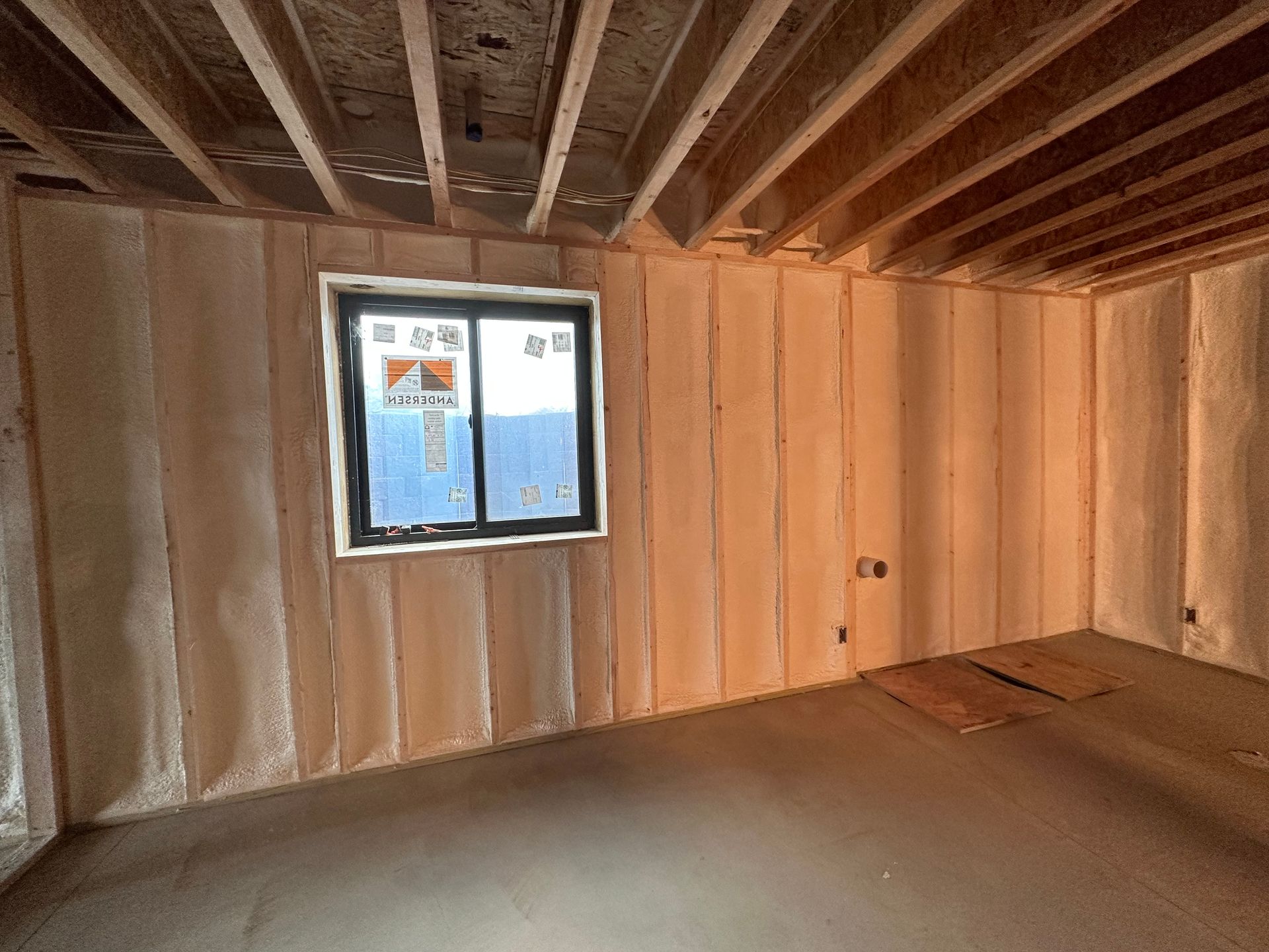 Bare unfinished room with exposed wooden studs, ceiling joists, and a small window showing blue sky.