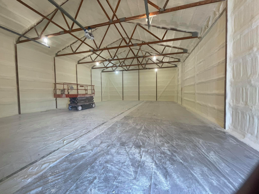 Empty industrial warehouse with white walls, exposed roof trusses, and a scissor lift on the left.