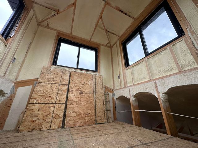 Unfinished attic room with exposed insulation, plywood walls, windows, and built-in cubbies.