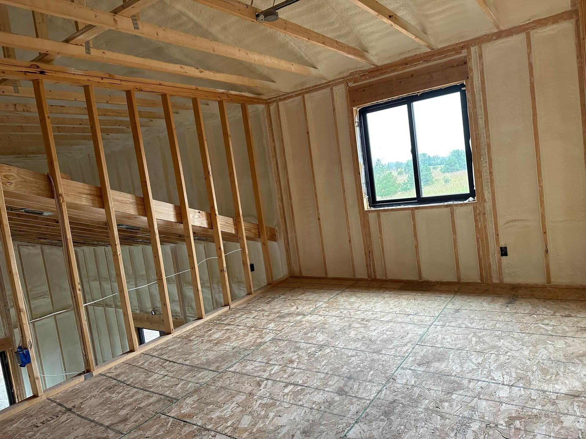 Unfinished attic room with exposed framing, insulation, plywood floor, and a small window.