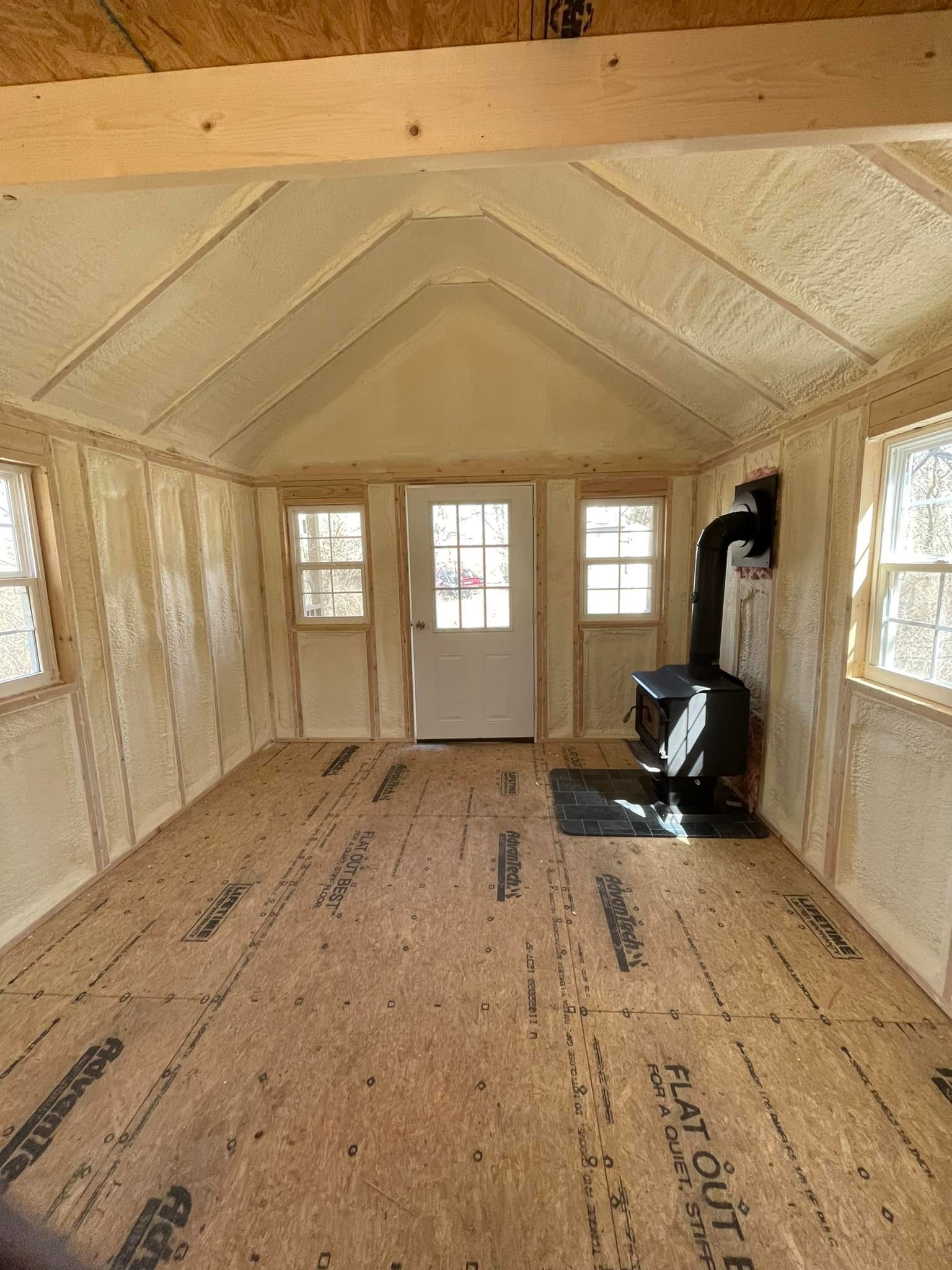 Bare insulated room with plywood floor, white door, small windows, and a wood stove on the right