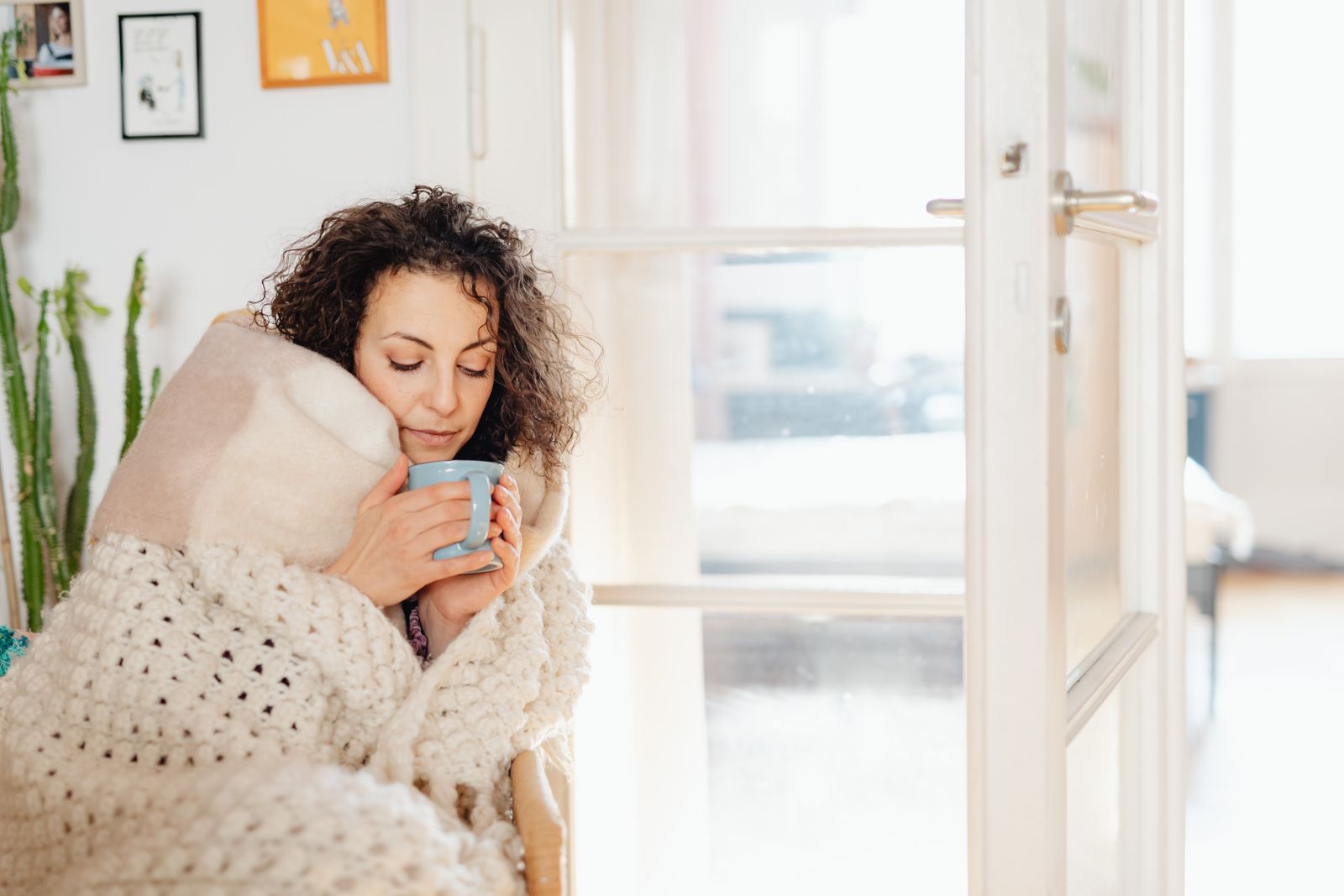 Woman wrapped in a blanket holding a warm mug inside a chilly, drafty home