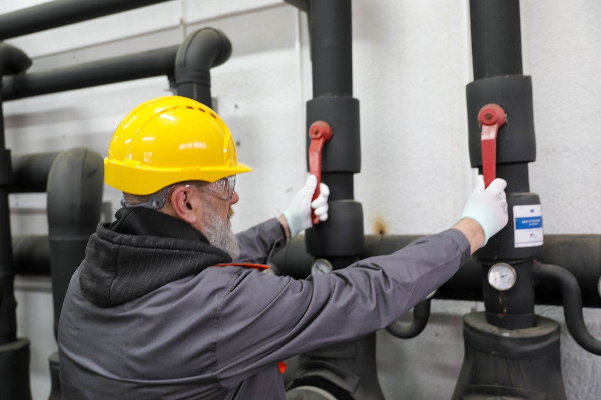 A man in a hard hat & safety glass shuts off a valve on pipes in a commercial plumbing facility.