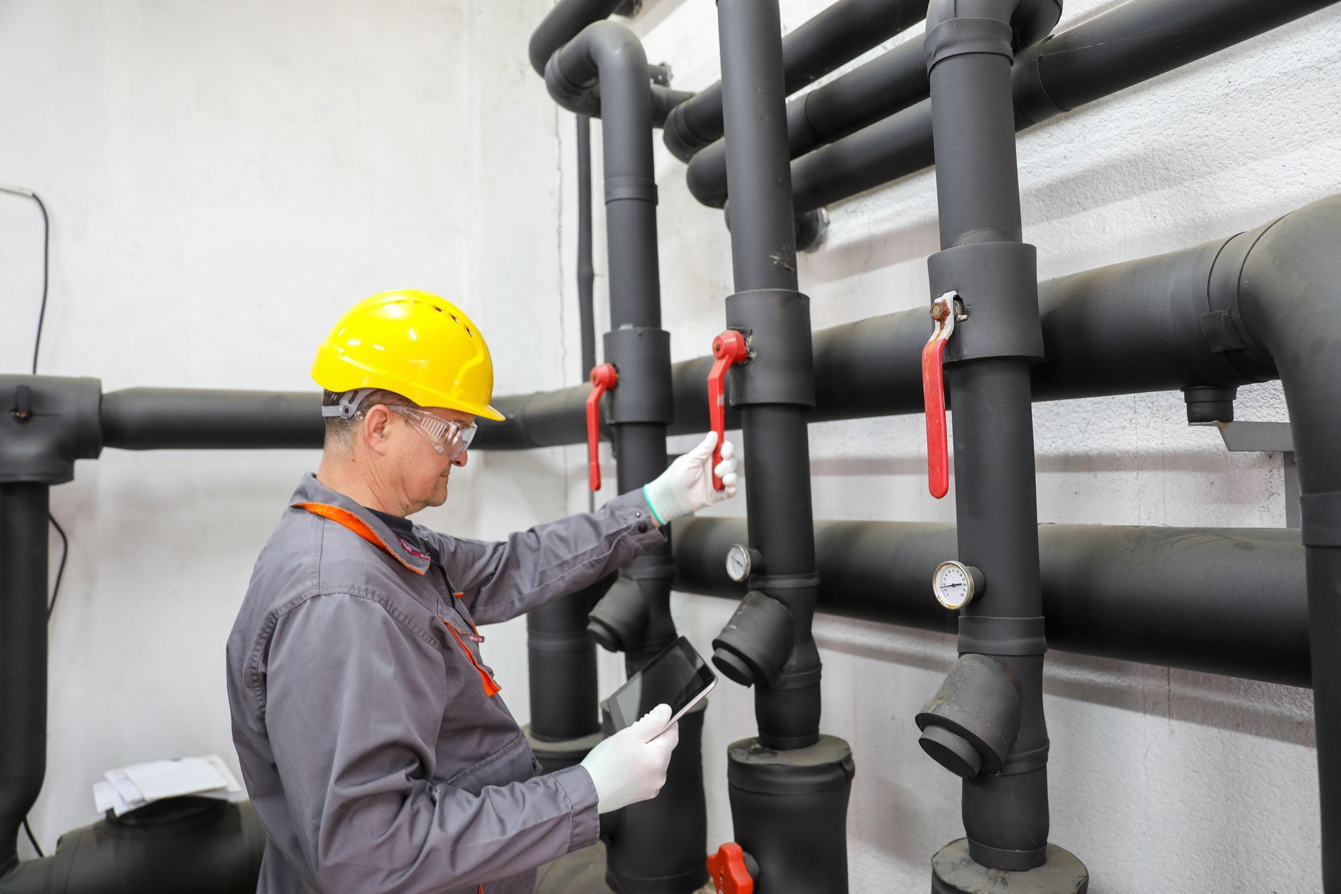 A man in a hard hat and safety glasses inspects pipes in a commercial plumbing facility.