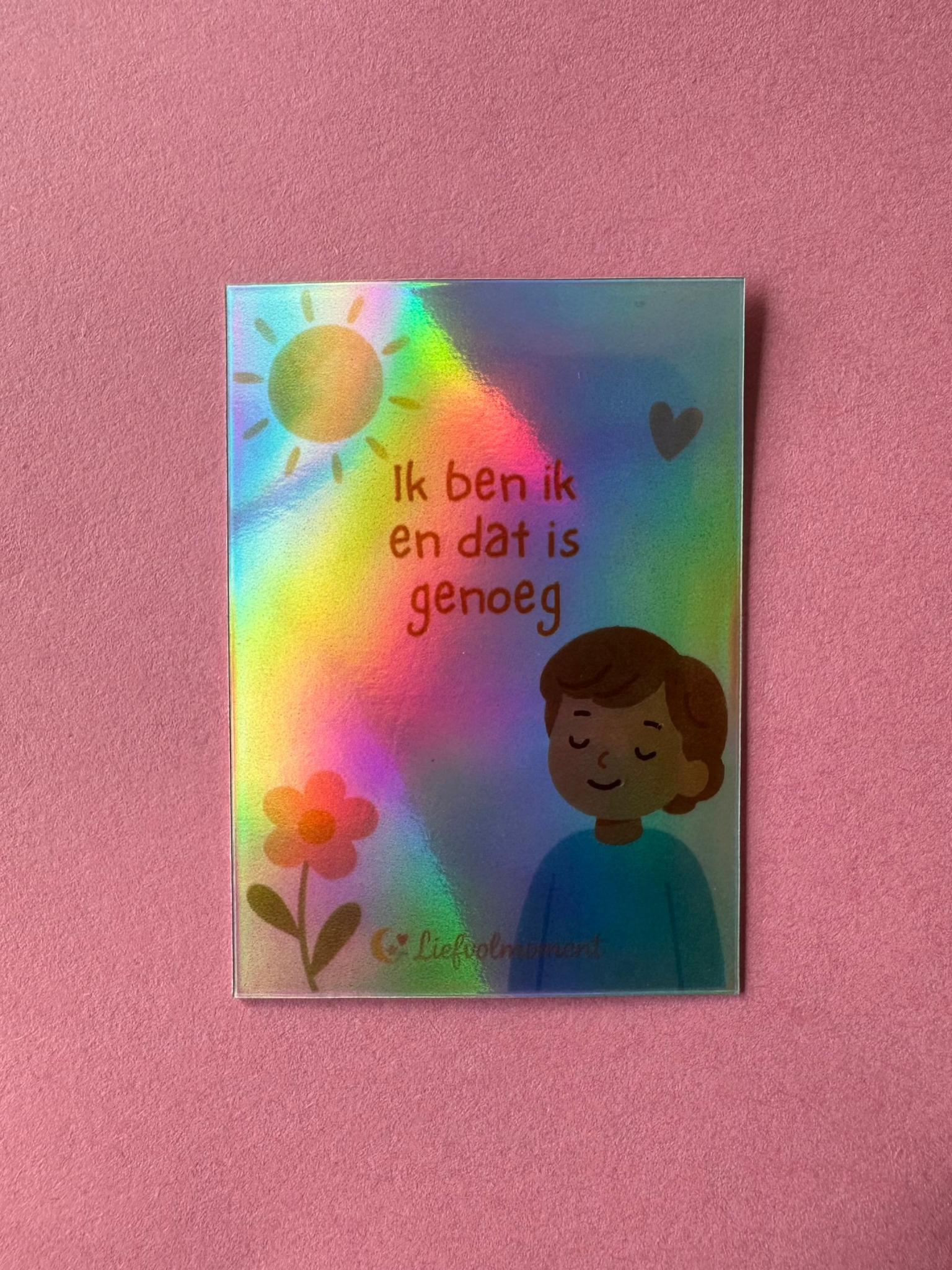 Een rechthoekige kaart met een holografische afwerking toont een persoon met een serene uitdrukking, een bloem, een zon en een hart.