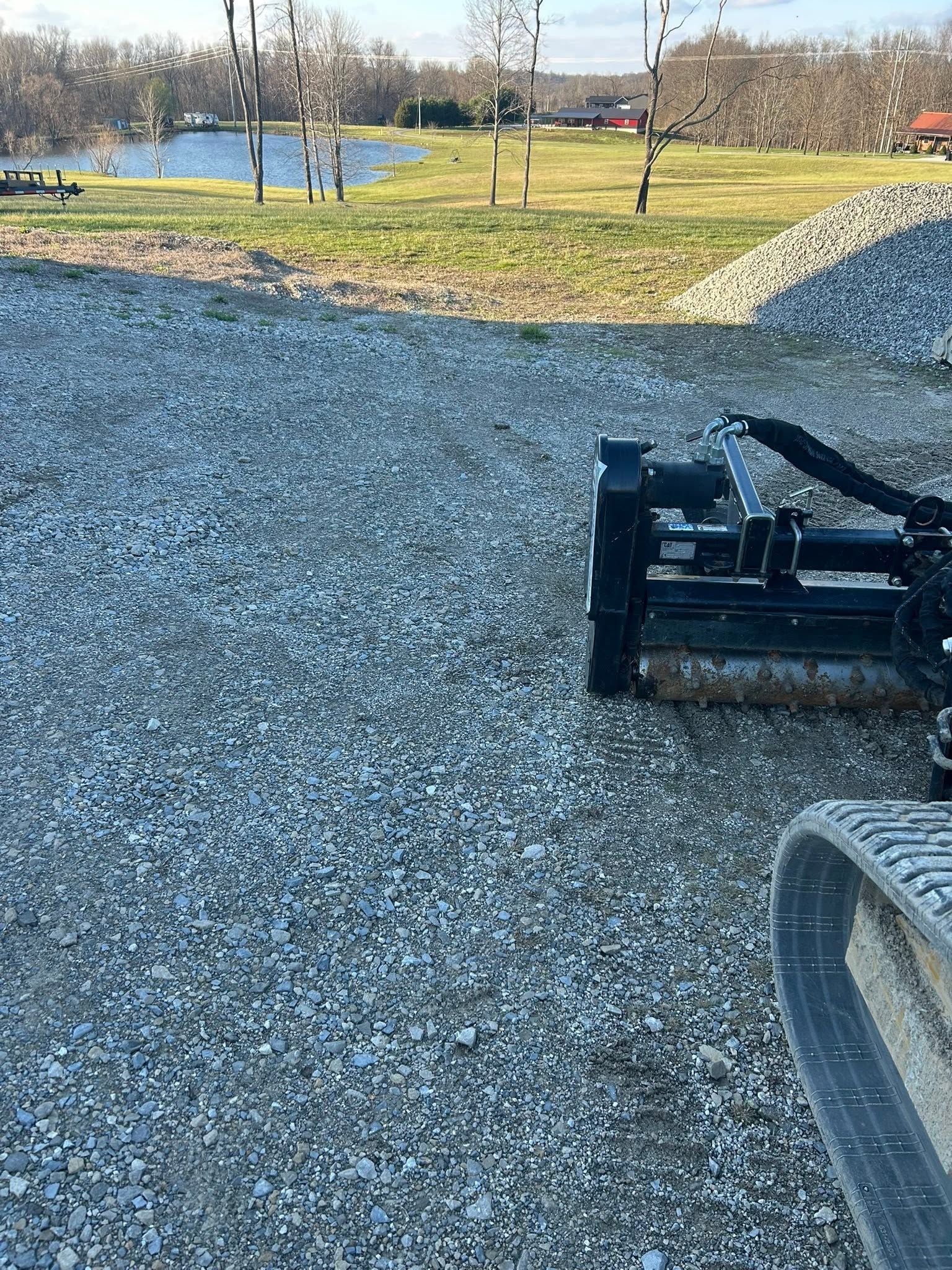 Gravel-covered path beside a pond and grassy park, with a black utility attachment in the foreground.