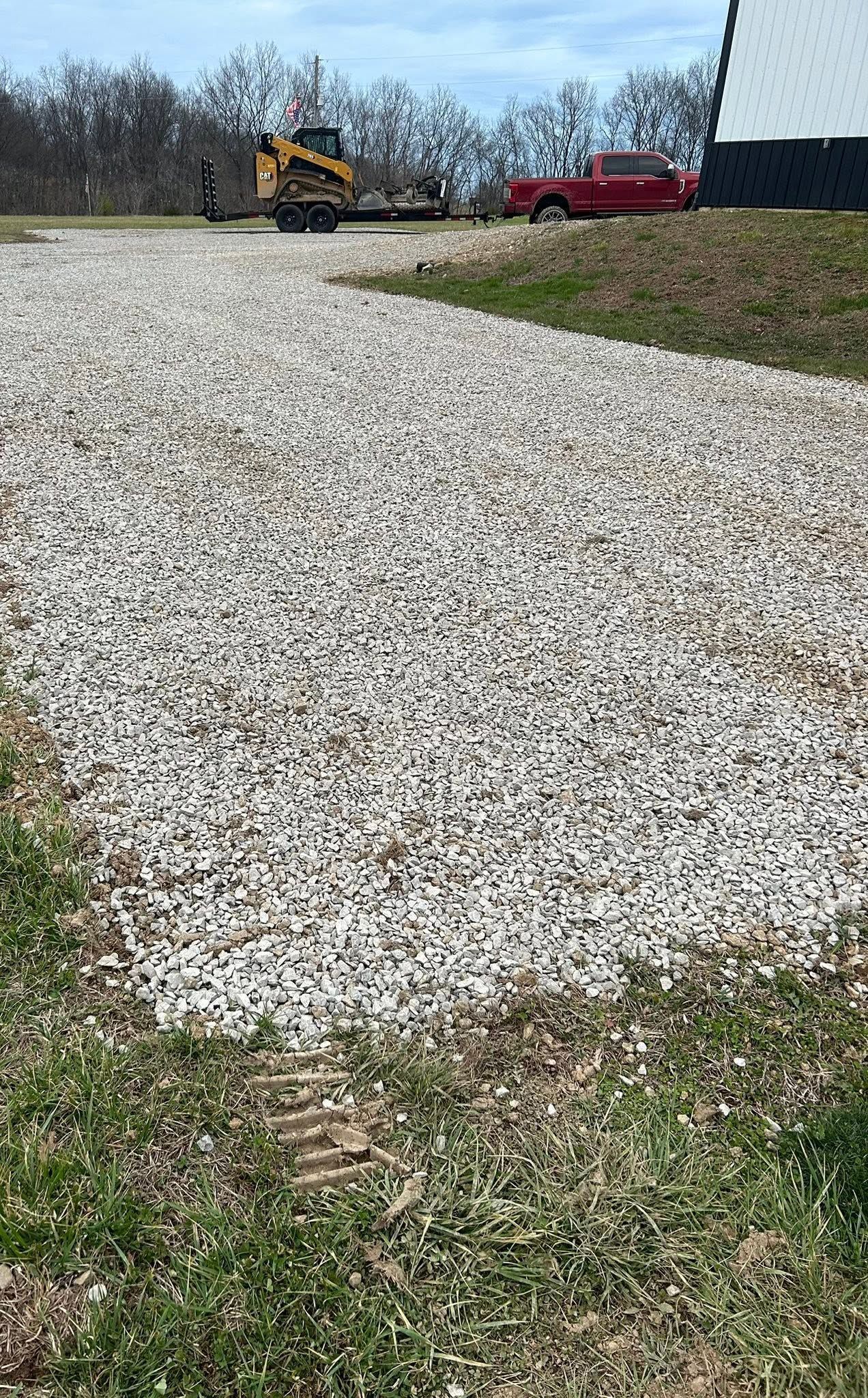 Gravel driveway leading to a yellow excavator and red pickup on a grassy lot