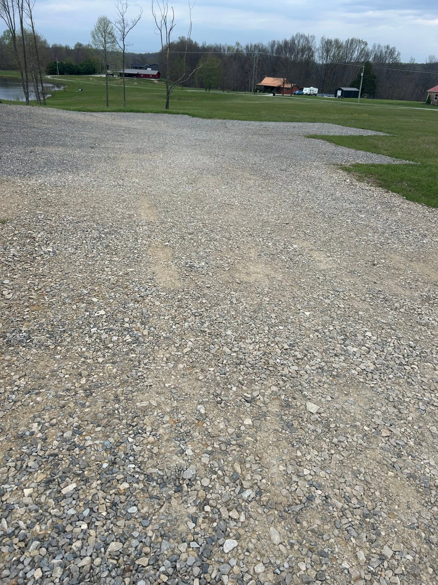 Gravel lot beside a grassy field with trees and a few houses in the distance.