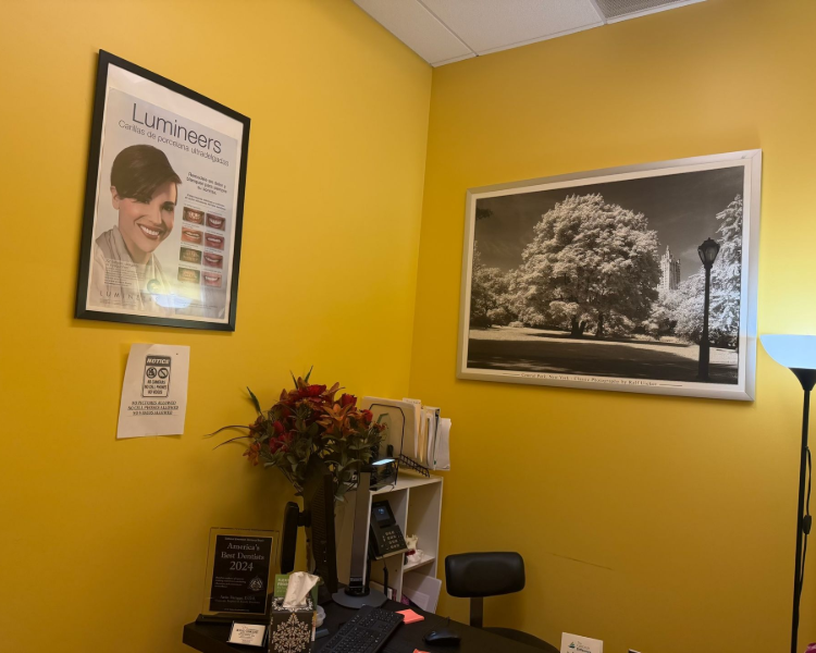 Small office with yellow walls, desk clutter, laptop, framed portrait, and sunset tree wall art.