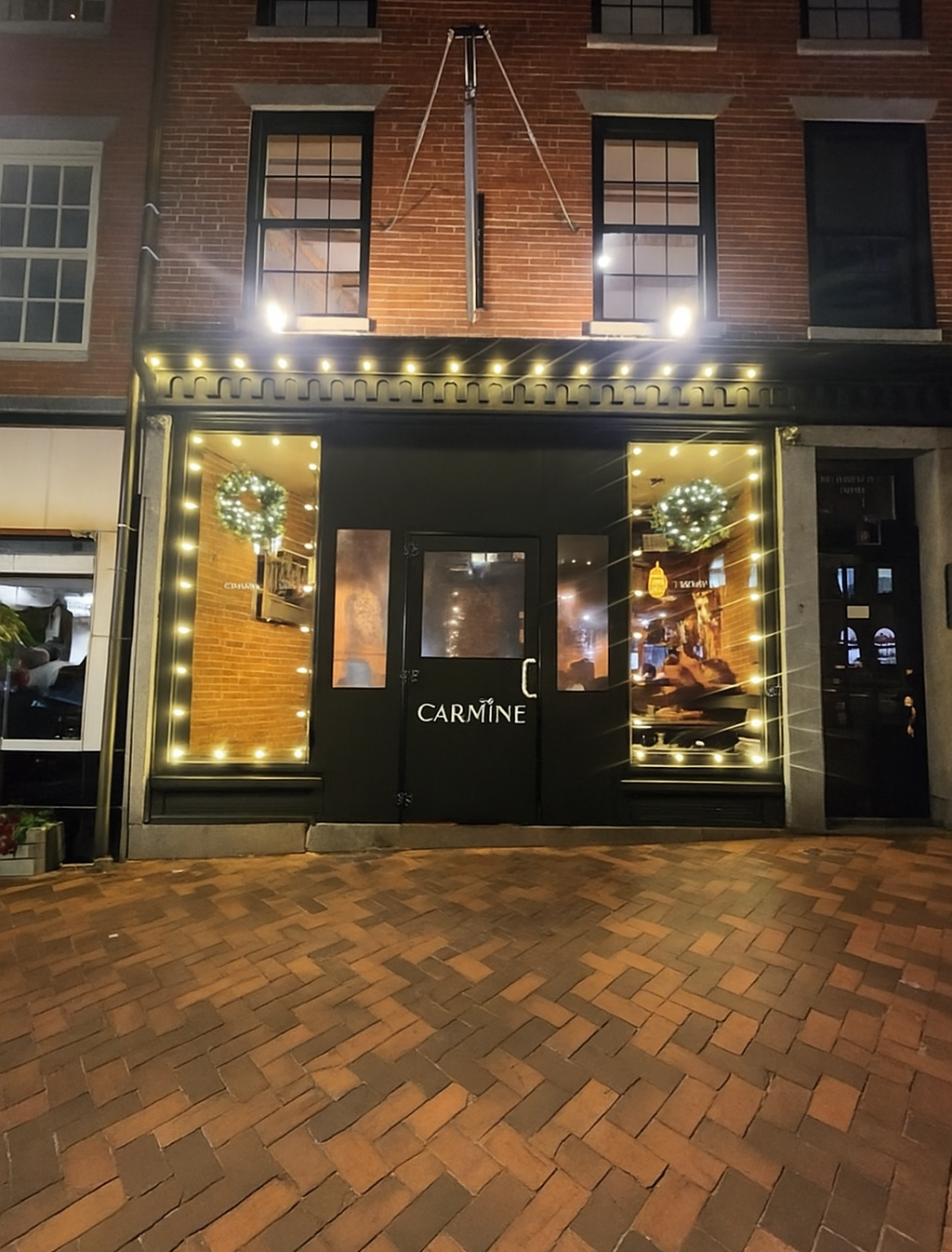 Storefront at night with lit holiday wreaths, gold window displays, and a brick walkway.