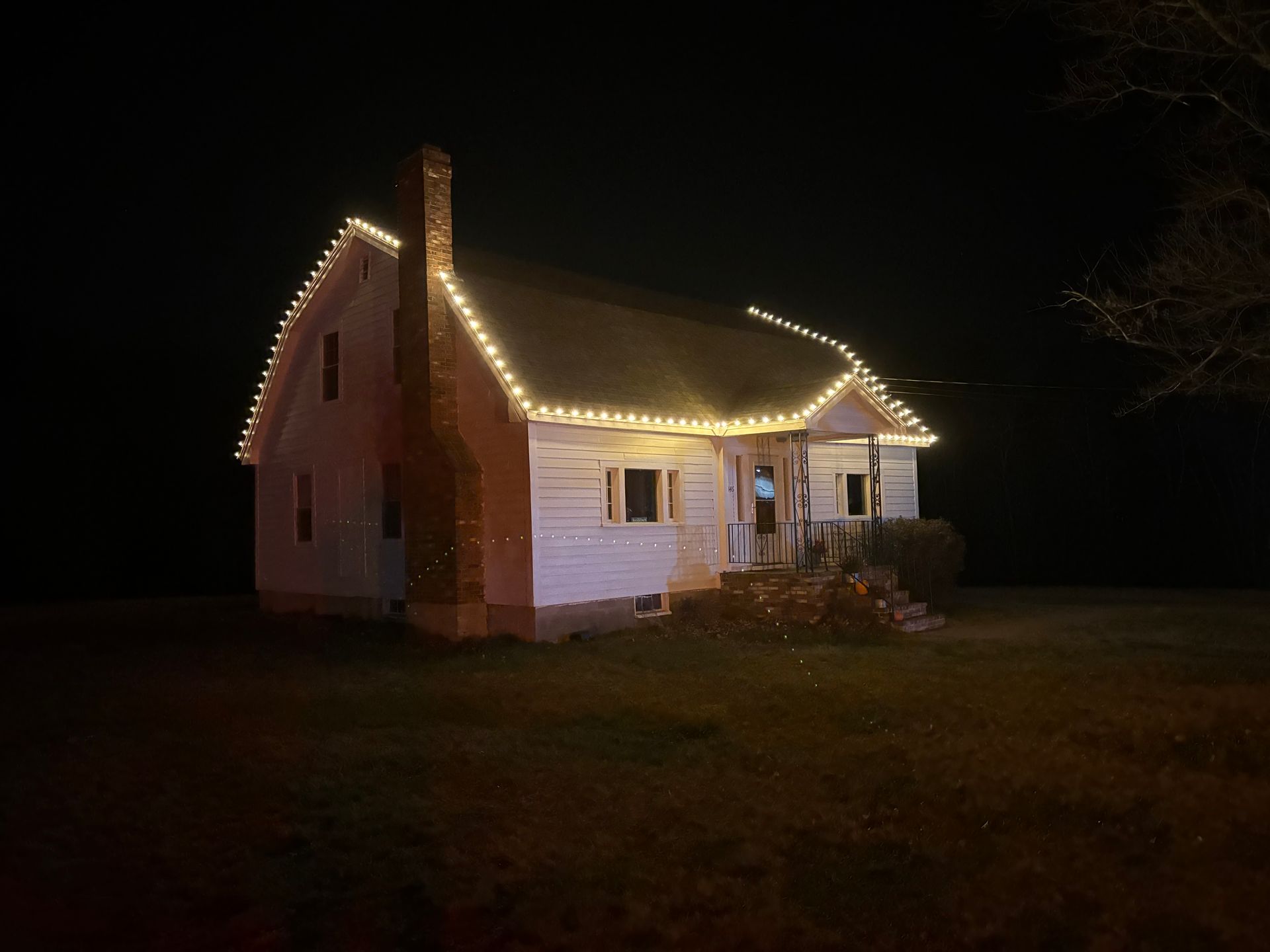 Small white house at night outlined with warm holiday lights, standing alone on a dark lawn.