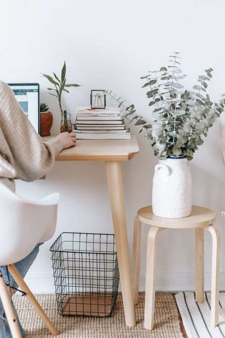 A cozy workspace with a laptop, stacked books, a small plant, and a white stool with a vase of greenery.