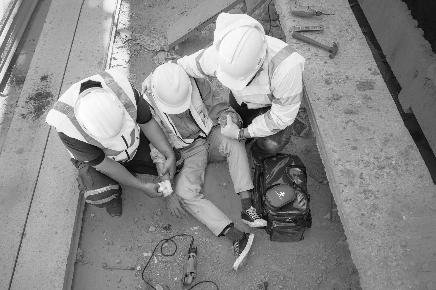 Two construction workers in high-visibility vests and hard hats provide medical assistance to an injured person.