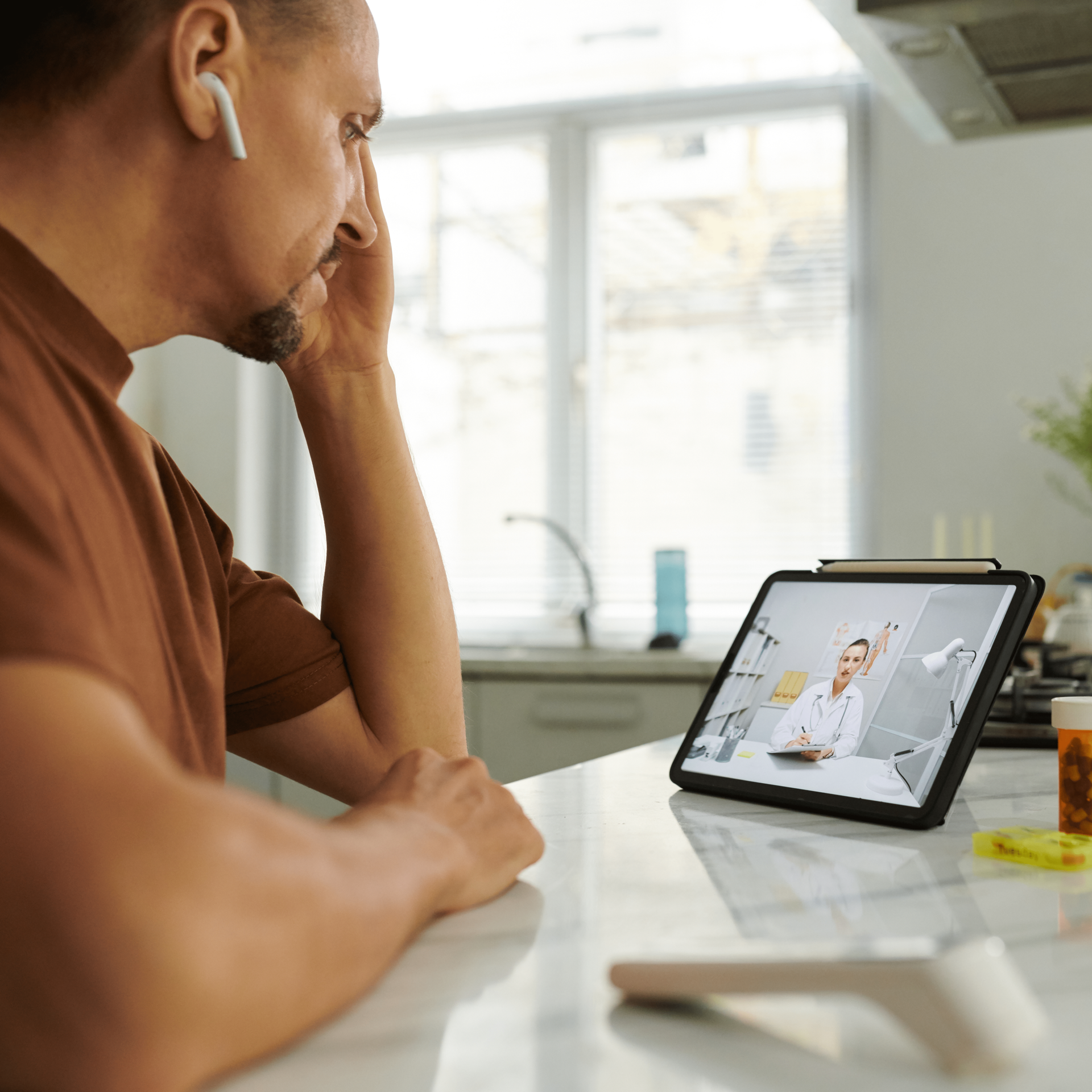 Man on video call with doctor on tablet, hand on forehead, prescription pills visible.