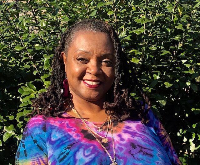Woman with locs smiles, wearing colorful tie-dye top, outdoors in front of green foliage.