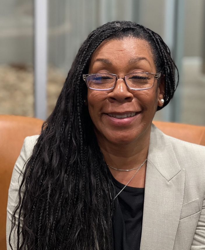 Woman with long braided hair wearing glasses and a blazer smiles, sitting indoors.