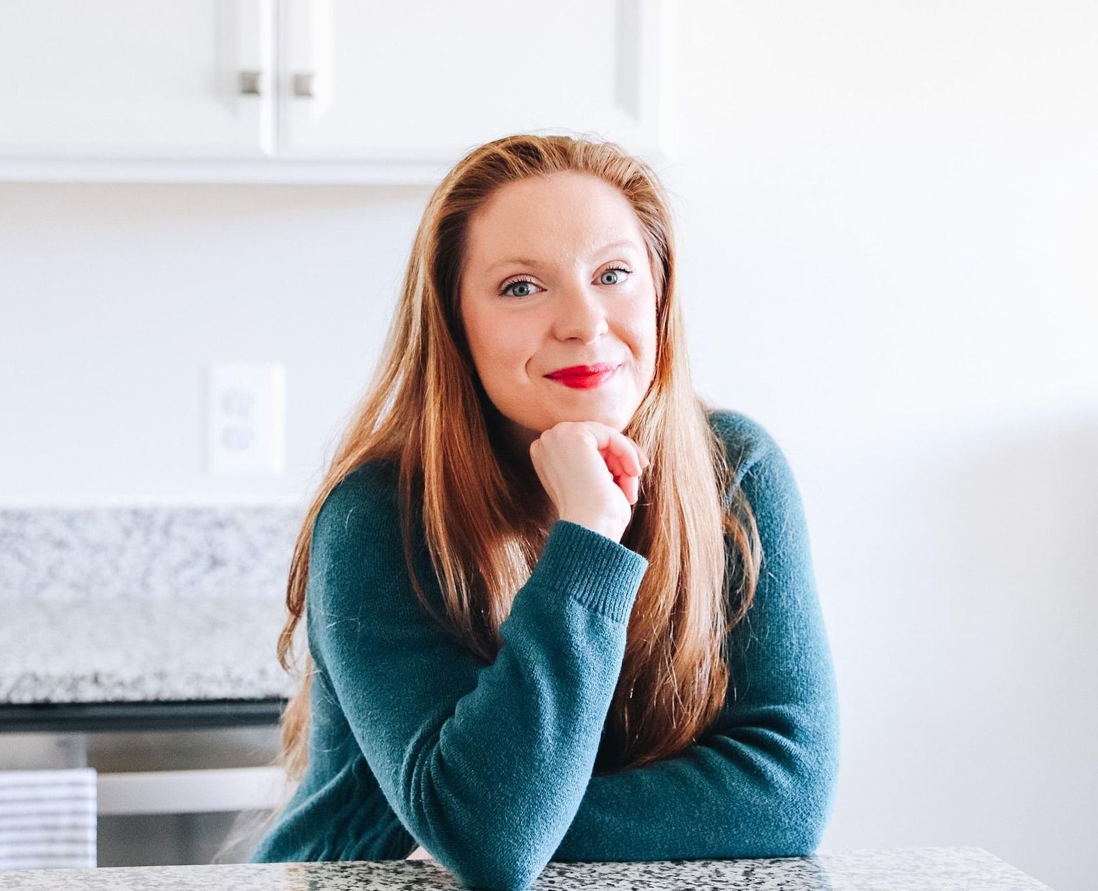 Woman with red lipstick, hand on chin, smiles at the camera, leaning on a countertop in a kitchen.             