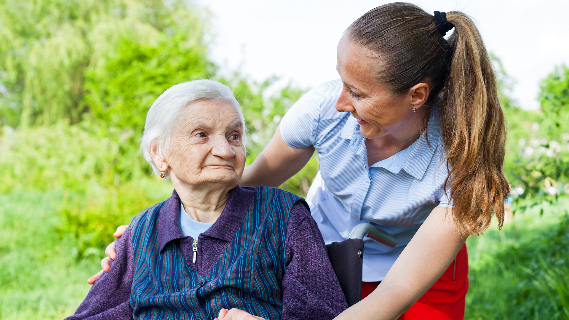 Woman assisting an elderly person in a wheelchair outdoors. Green background. Both smiling.