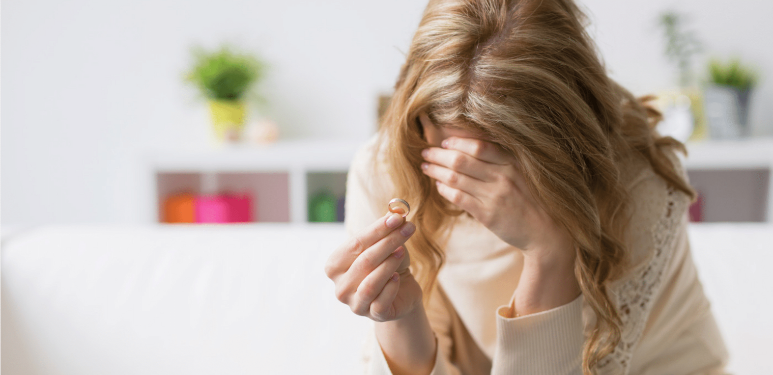 A woman holding a ring, covering her face with her hand. She appears sad, sitting on a couch.