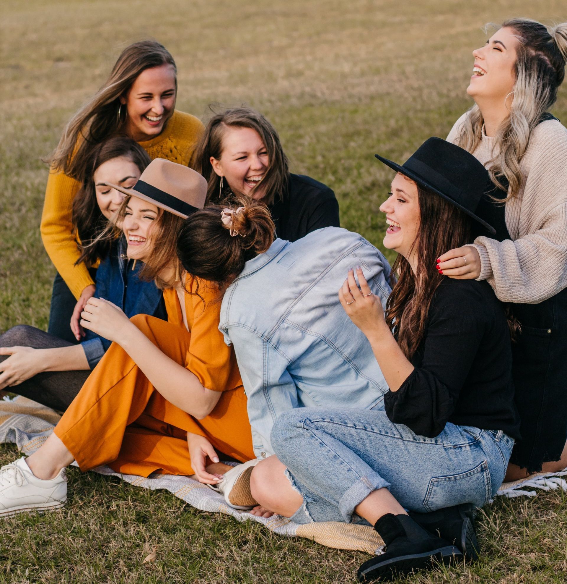 Group of women laughing and sitting on a blanket in a grassy field.