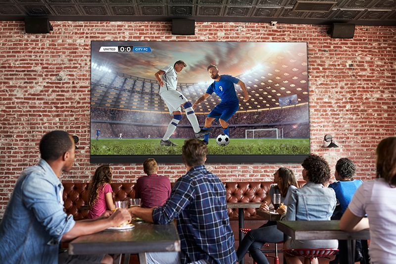 A group of people are sitting at tables watching a soccer game on a large screen.