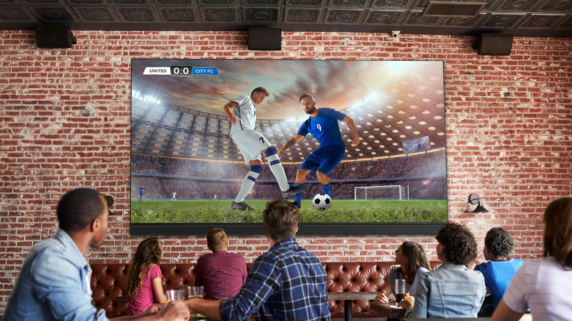 A group of people are sitting at tables watching a soccer game on a large screen.