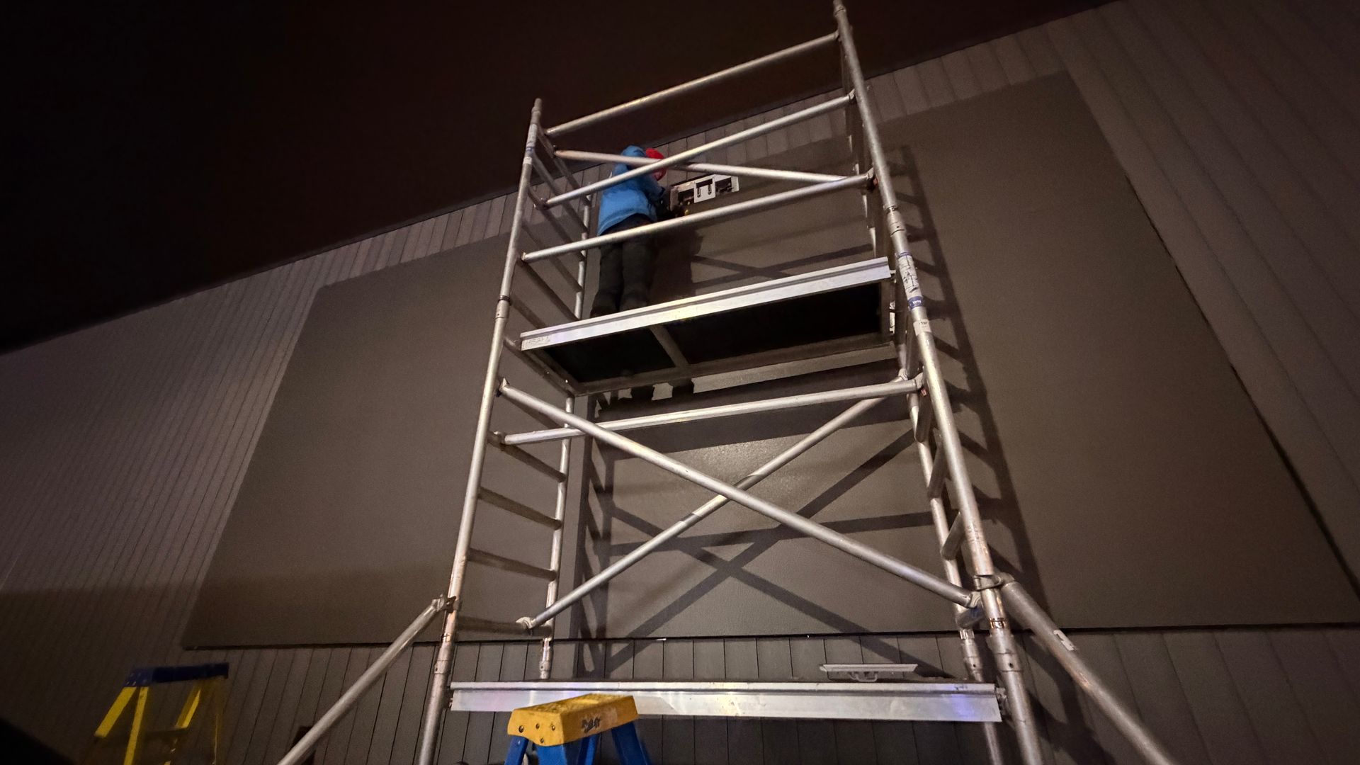 Person on scaffold installing panel on wall. Dark interior, neutral colors.