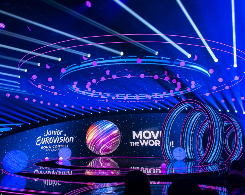 Stage set for Junior Eurovision Song Contest, with blue and pink lights, circular design, and logo.
