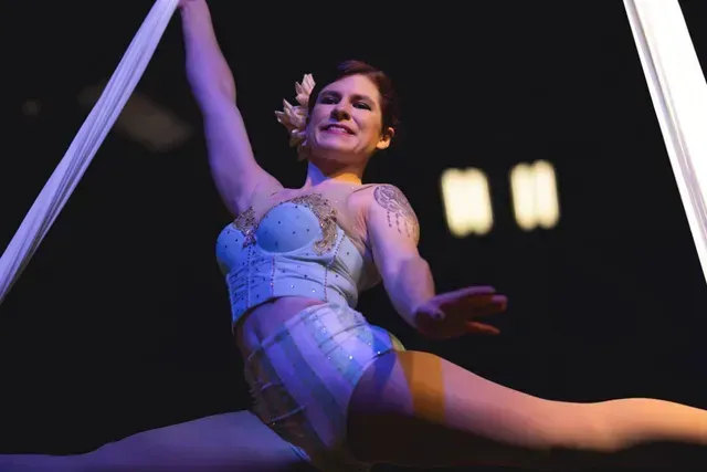 An aerialist in a light blue costume performs a split while suspended by white fabric silks against a dark background.