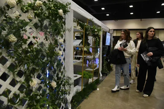 People walk past a floral-decorated trellis and display shelves at a trade show or indoor event.