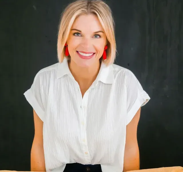 A person with blonde hair in a white shirt and red earrings smiles against a dark background.