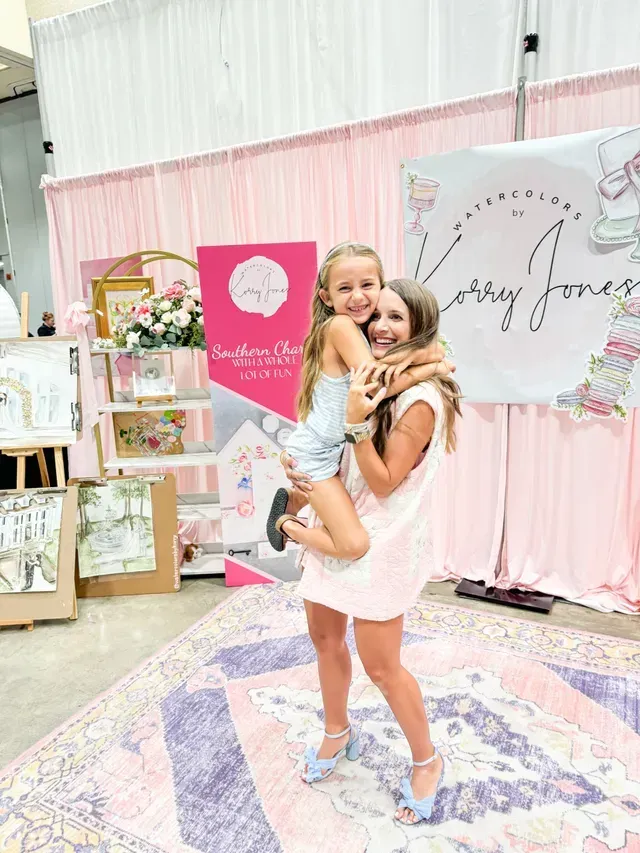 A woman holding a child in her arms, both smiling, at a decorated event booth with pink drapes and watercolor signs.