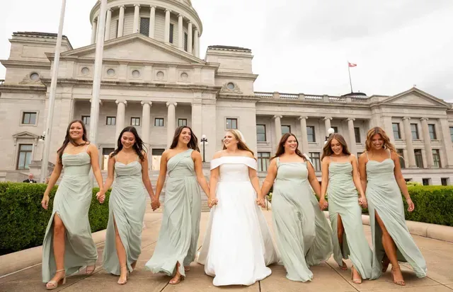 A bride in a white gown and six bridesmaids in sage green dresses walk holding hands in front of a stone capitol building.