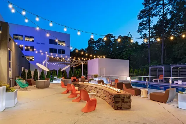 An outdoor patio at dusk with a long stone fire pit, modern orange seating, string lights, and a swimming pool.
