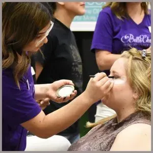 A person in a purple shirt applies eye makeup to another person using a small brush.