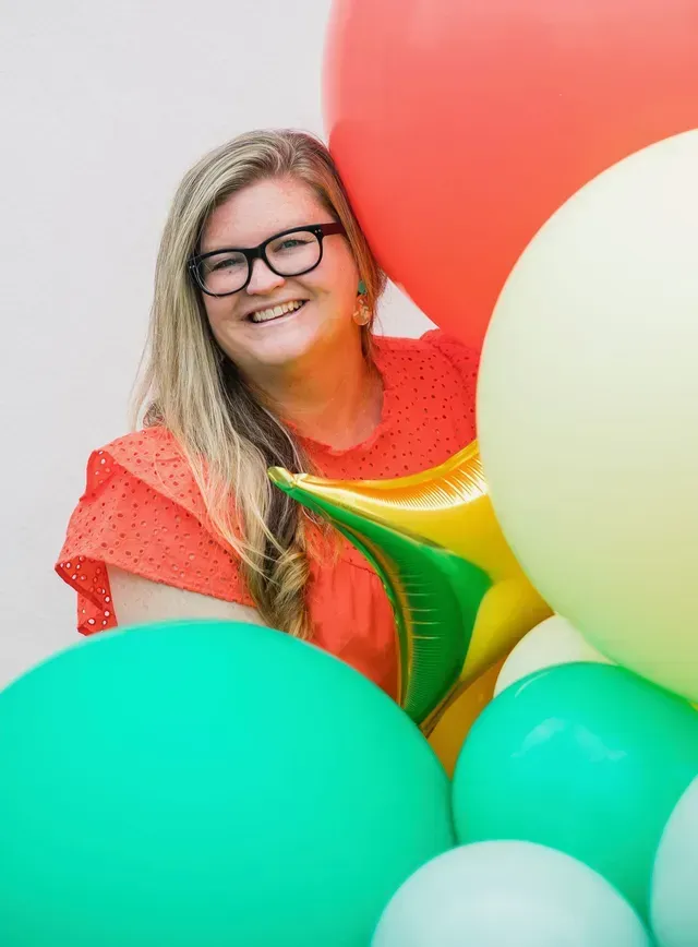 A smiling person with glasses wearing an orange top, surrounded by large, colorful balloons in a bright setting.